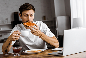 emotional young freelancer eating toast with jam and coffee and looking at laptop screen at home