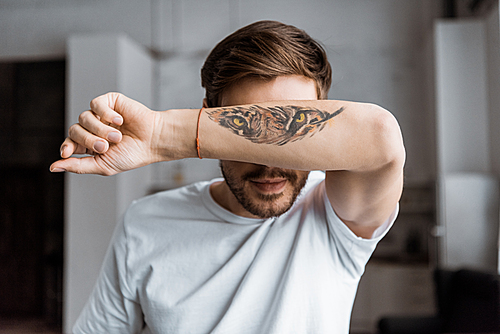 handsome young man covering face with arm with tattooed tiger eyes at home