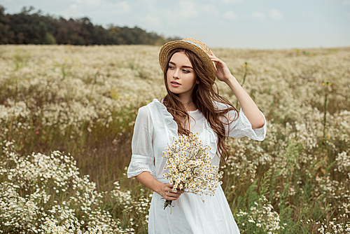 portrait of pretty pensive woman in white dress with bouquet of wild camomile flowers on meadow