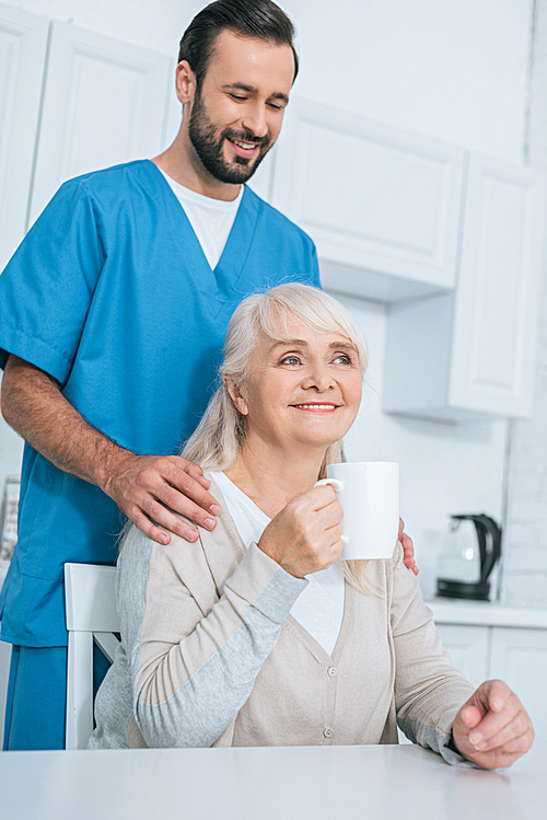 smiling male caregiver looking at senior woman drinking tea in kitchen