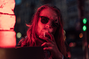 handsome young man in sunglasses smoking cigarette under red light on street
