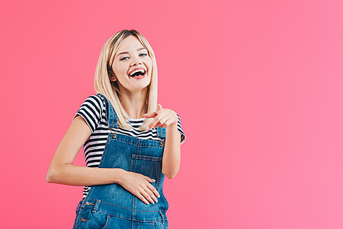 laughing girl in denim overall pointing on camera isolated on pink