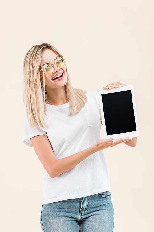 cheerful young woman in stylish eyeglasses showing digital tablet with blank screen isolated on beige