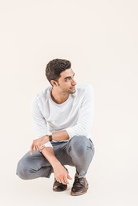 handsome smiling young man crouching and looking away isolated on beige
