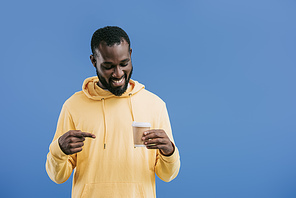 smiling young african american man pointing by finger at disposable coffee cup isolated on blue background