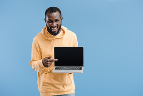 laughing young african american man pointing by finger at laptop with blank screen isolated on blue background