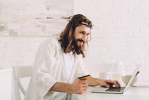 cheerful Jesus holding credit card and doing online shopping at table with laptop in kitchen at home
