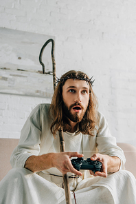 emotional Jesus with wooden staff playing video game by joystick on sofa at home