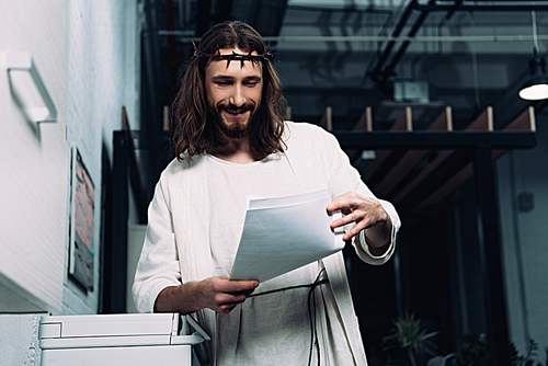 selective focus of smiling Jesus in crown of thorns and robe checking ...
