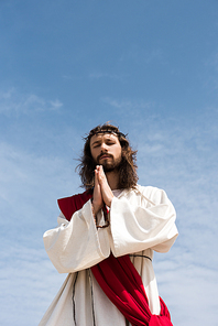 low angle view of Jesus in robe, red sash and crown of thorns holding rosary and praying with closed eyes against blue sky