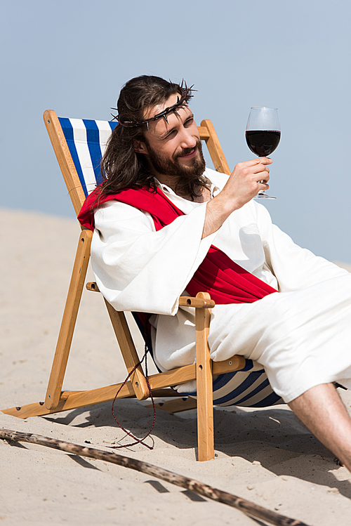 cheerful Jesus in robe and red sash resting on sun lounger and looking at glass of red wine in desert