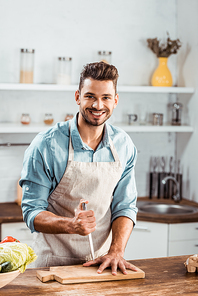 handsome young man in apron holding knife and smiling at camera in kitchen