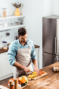 high angle view of young man in apron cutting fresh peppers in kitchen