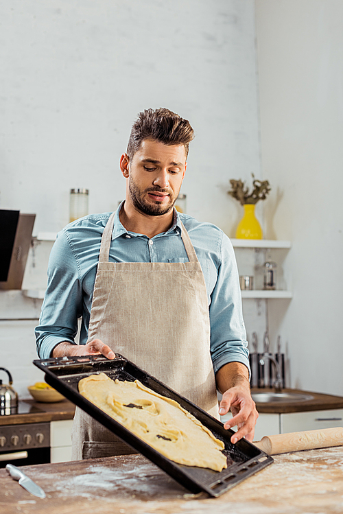 frustrated young man in apron looking at spoiled dough on baking tray