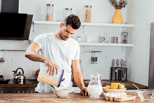 young man holding container with corn flakes and preparing breakfast in kitchen