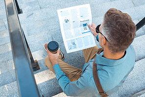 high angle view of man with paper cup sitting on stairs and reading newspaper