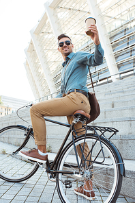 low angle view of smiling man sitting on bicycle and holding disposable coffee cup