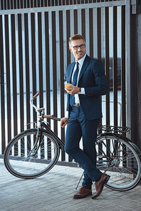 smiling middle aged businessman leaning at bicycle and holding hamburger on street