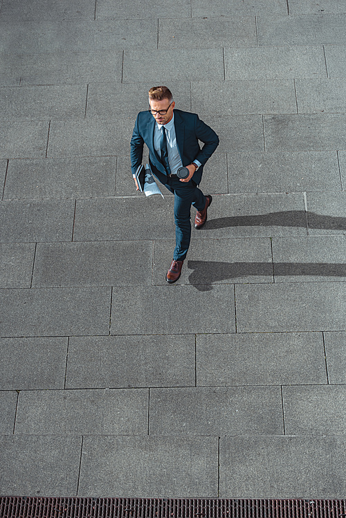 high angle view of businessman with paper cup and newspaper walking on street