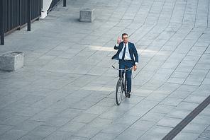 high angle view of middle aged businessman riding bicycle and showing victory sign