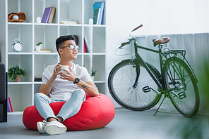 handsome asian man sitting on bean bag chair with cup of tea and looking away at home