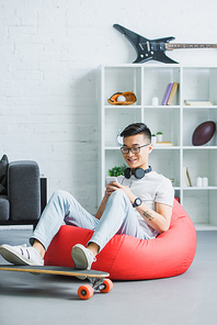 smiling young asian man sitting in bean bag chair and using smartphone at home
