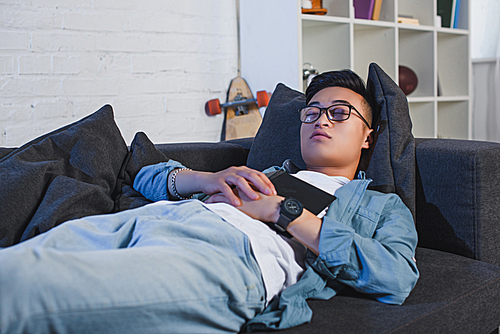young asian man in eyeglasses holding book and sleeping on couch