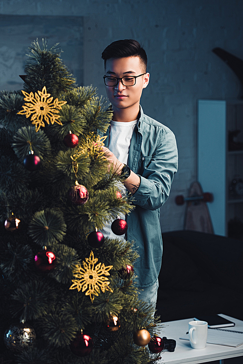 young asian man in eyeglasses decorating christmas tree at home