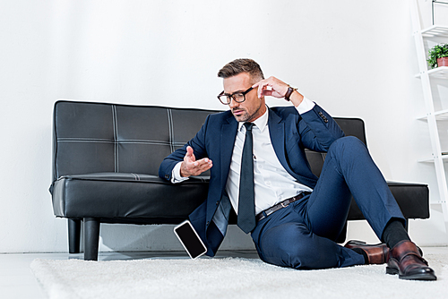 handsome businessman in suit sitting on carpet near sofa and looking at smartphone with blank screen