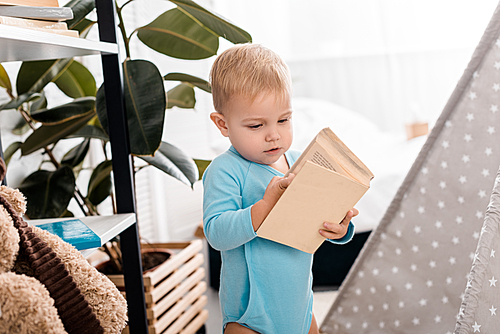 cute toddler boy in blue bodysuit holding book in bedroom