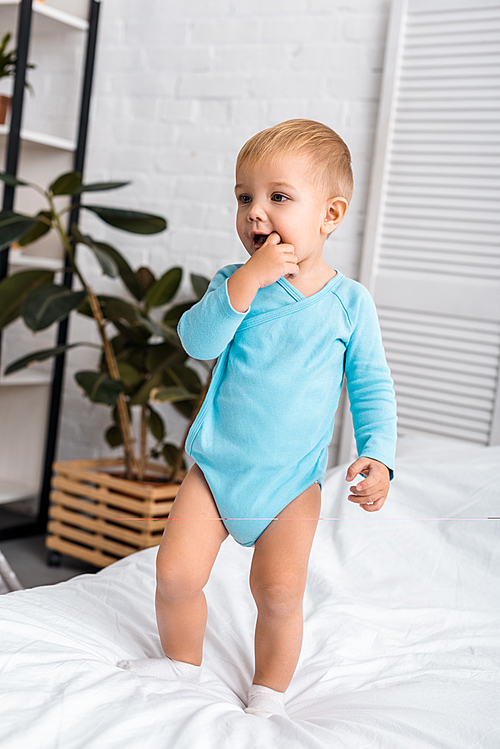 cute baby putting finger in mouth and standing on bed in light room
