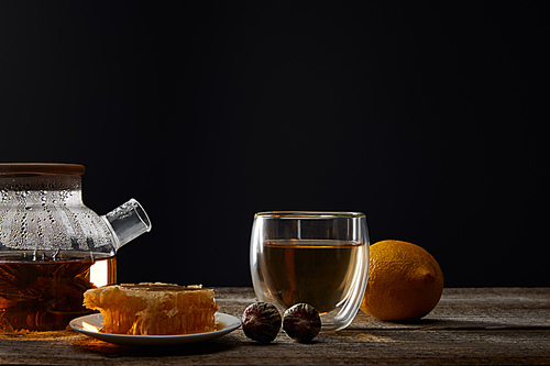 transparent teapot with blooming tea, glass, lemon and honeycomb on wooden table isolated on black