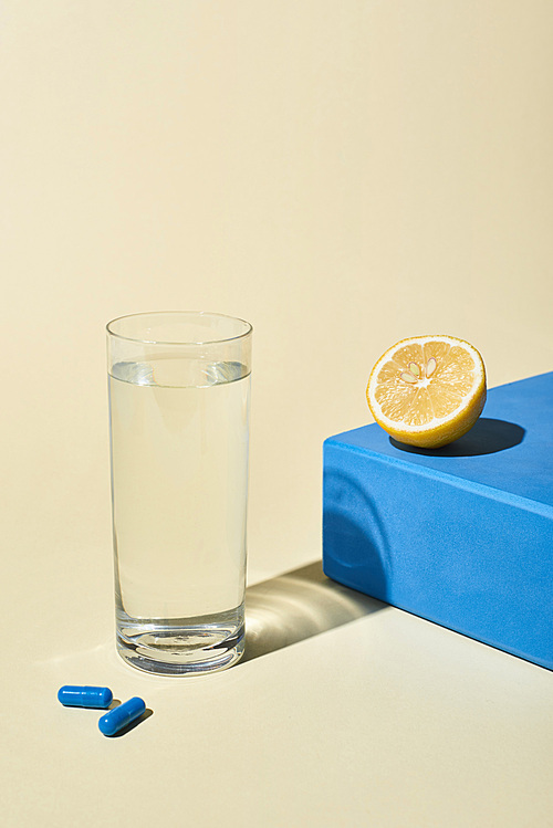 glass of water, blue pills and lemon half on blue cube on beige background