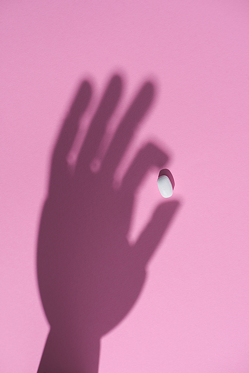 top view of shadow of hand holding pill on pink surface