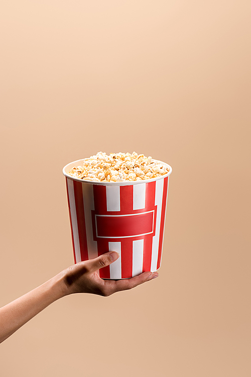 partial view of woman holding disposable bucket with popcorn isolated on beige