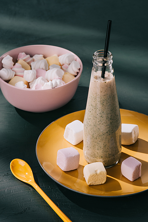 closeup shot of milkshake in bottle with  straw, bowl with marshmallows and spoon on tabletop