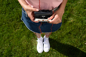 cropped shot of woman with earphones holding retro cassette player with audio cassette in hands while standing on green grass