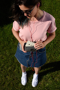 high angle view of stylish young woman in sunglasses and earphones with retro audio cassette and player in hands standing on green grass
