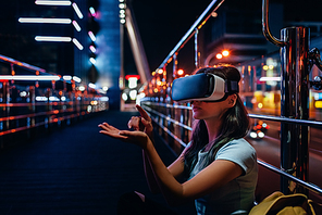 young woman in virtual reality headset sitting on street with night city on background