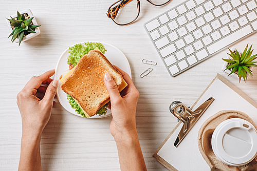 cropped image of businesswoman having lunch with sandwich and coffee in paper cup at table with computer keyboard in office