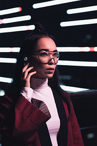 attractive asian girl in burgundy kimono and wireless earphone looking away on street with neon light in evening, city of future concept