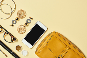 Top view of earrings, glasses, eyeshadow, smartphone, mascara, bracelets, cosmetic brush and bag on yellow background