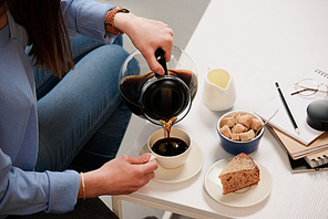 cropped shot of woman pouring coffee into cup with cane sugar and piece of cake on coffee table