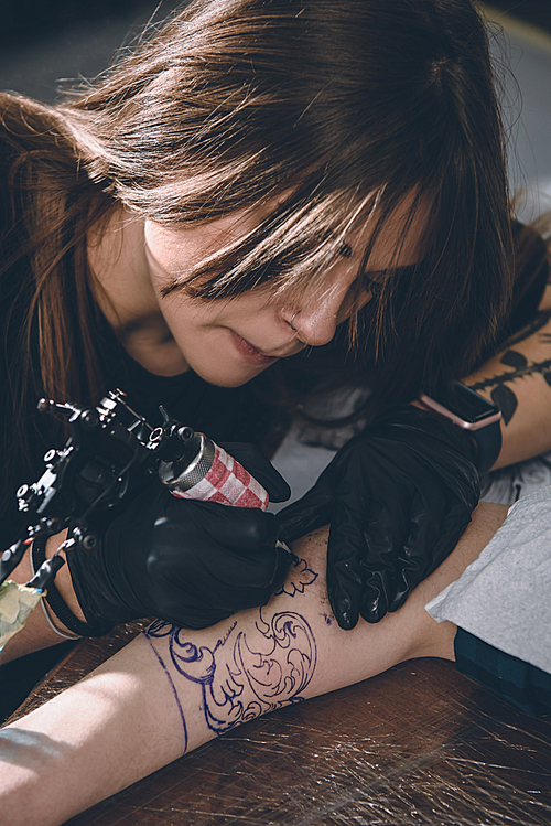 Concentrated female artist in gloves working on arm piece tattoo in studio