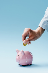 partial view of man putting coin into pink piggy bank isolated on blue