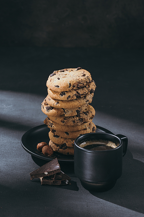 stack of chocolate-chip cookies on plate with cup of coffee on black surface