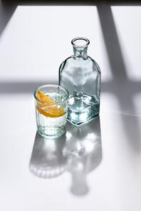 close up view of glass with lemon piece and bottle with water on white surface