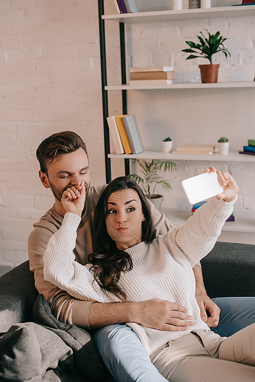 playful young couple taking selfie with smartphone on couch at home ...
