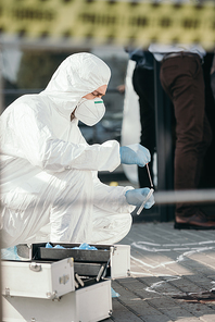 male criminologist in protective suit and latex gloves collecting blood sample in test tube at crime scene