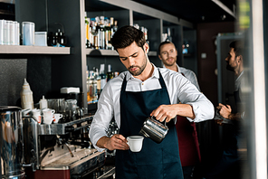 adult barista in apron pouring milk to coffee at workplace